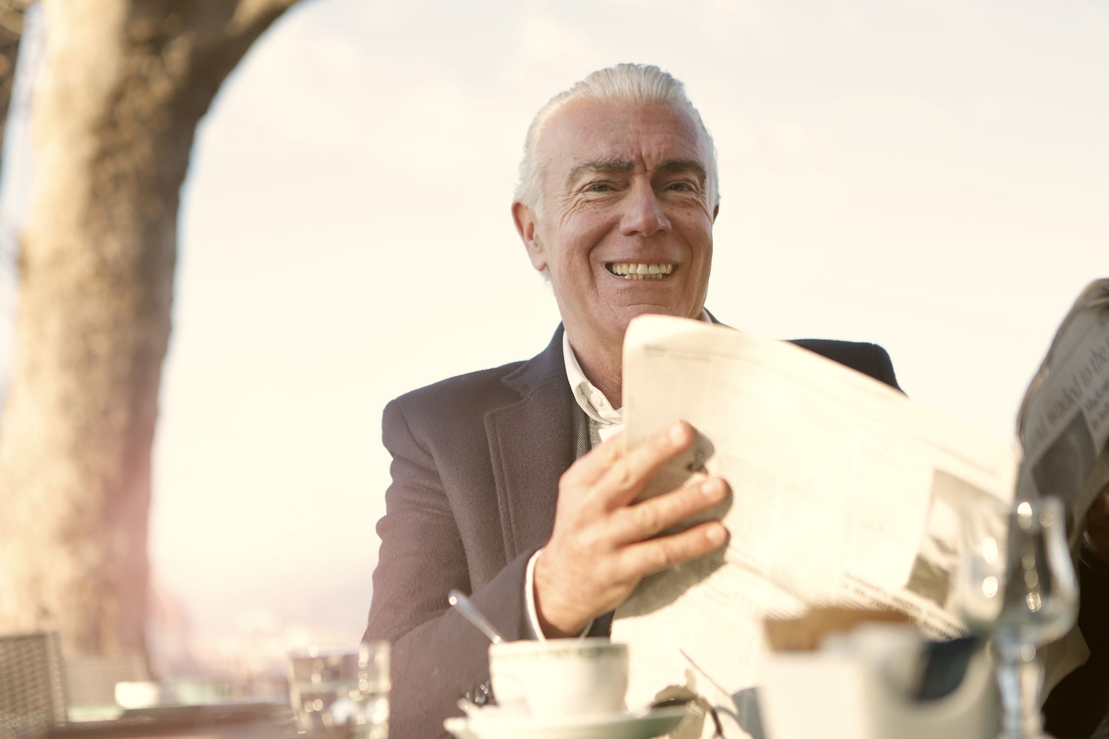 Man over 60 with white hair reading a newspaper at an outdoor cafe table in warm sunlight