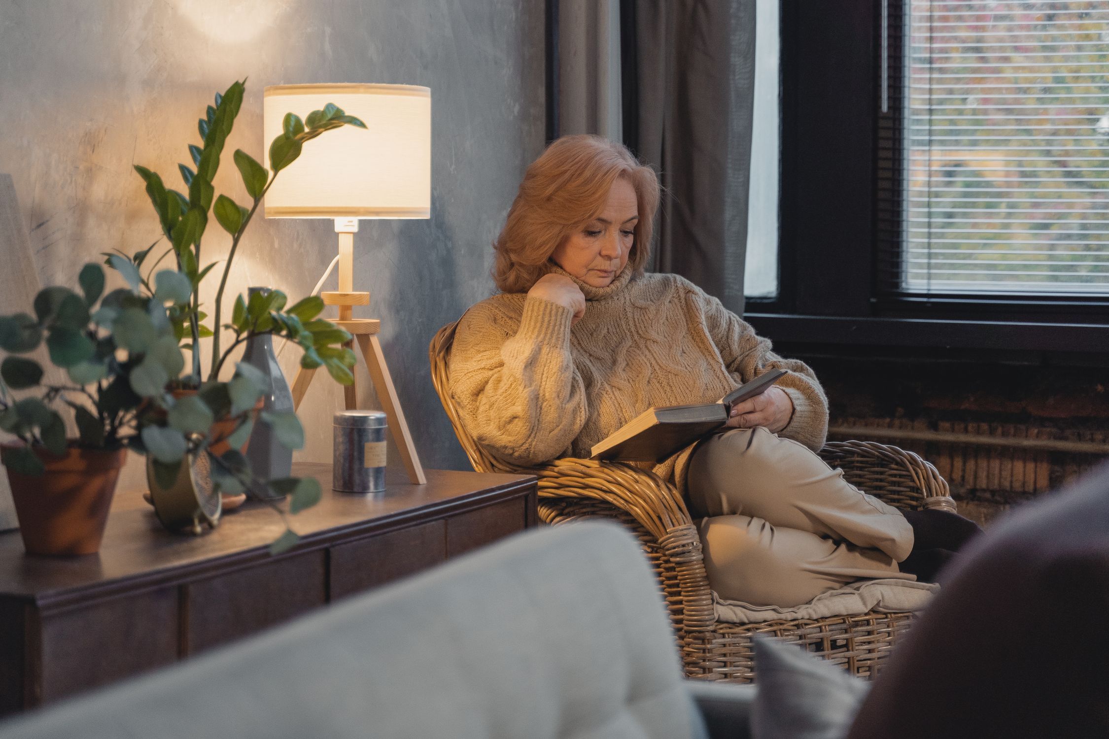 Woman over 50 reading a book in a wicker chair beside a warm lamp and houseplants