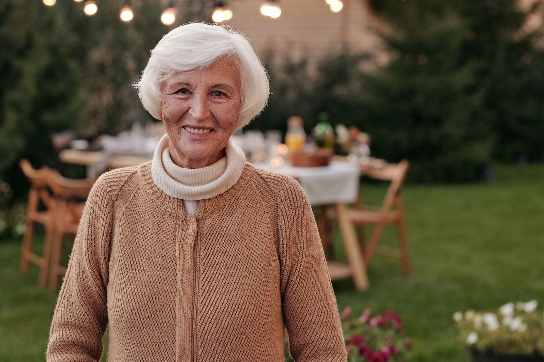 Woman over 60 with white hair smiling in a garden with string lights and a dinner table behind her