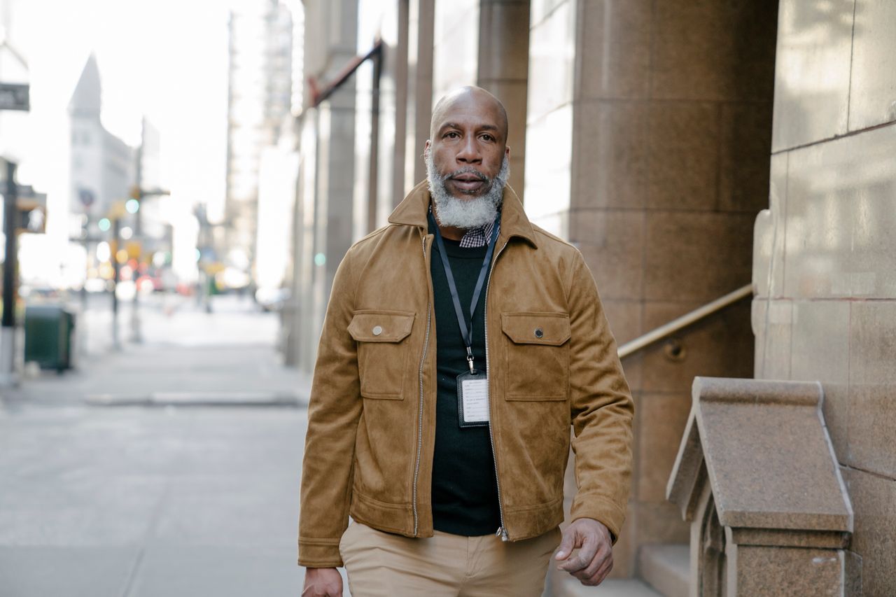 Man with gray beard in a tan jacket walking confidently on a city sidewalk