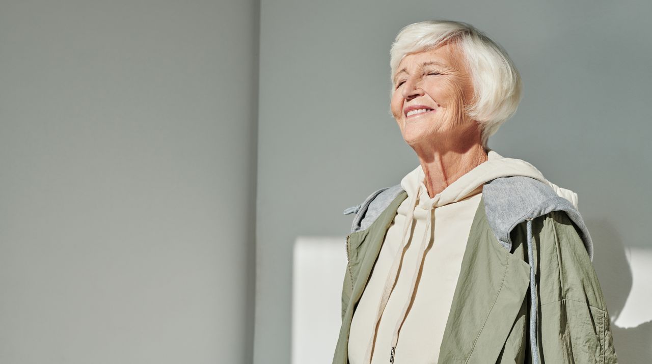 White-haired woman in an olive jacket smiling with eyes closed in warm sunlight