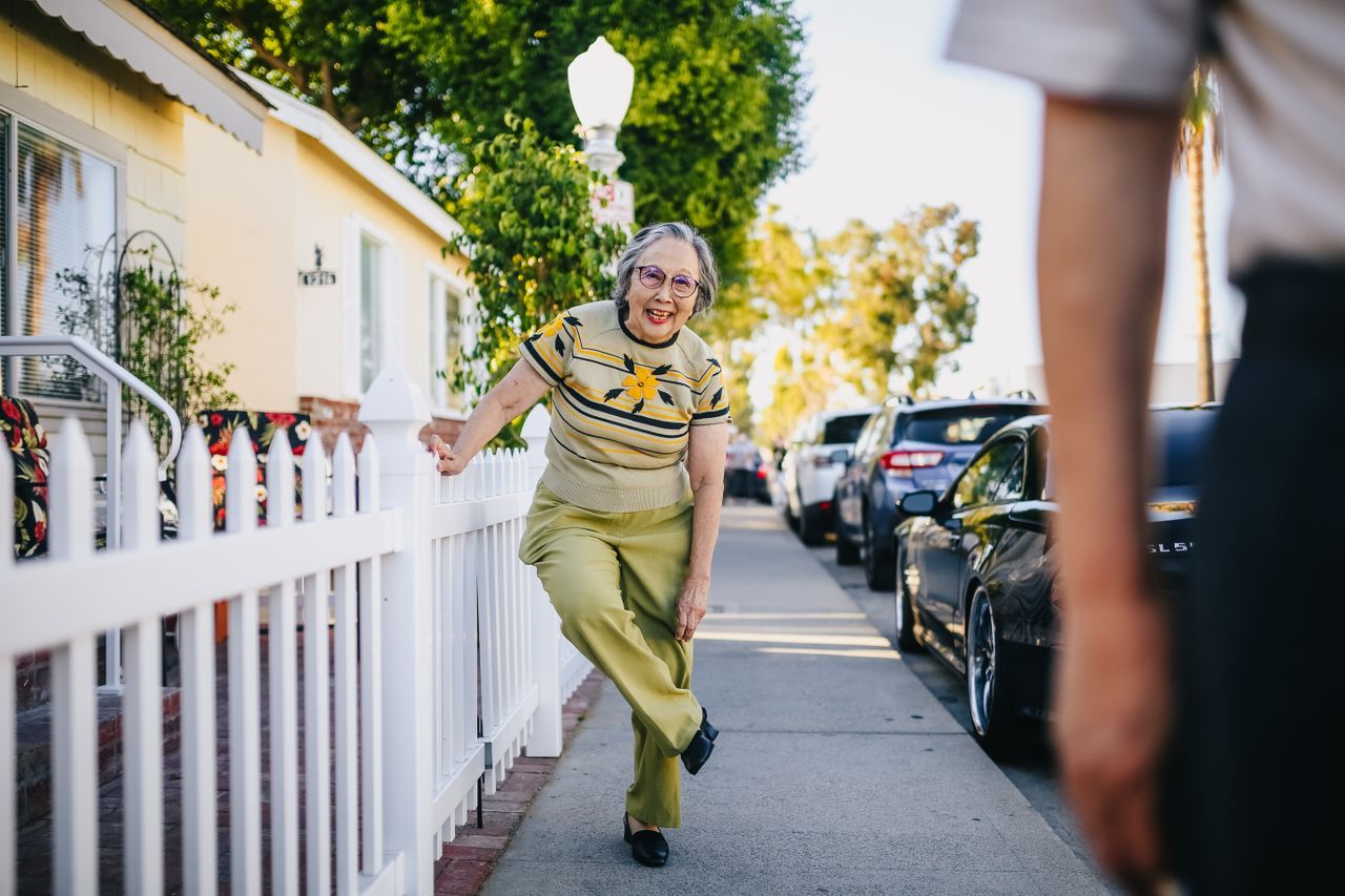 Senior woman on a sunny sidewalk leaning against a picket fence and smiling at a neighbour