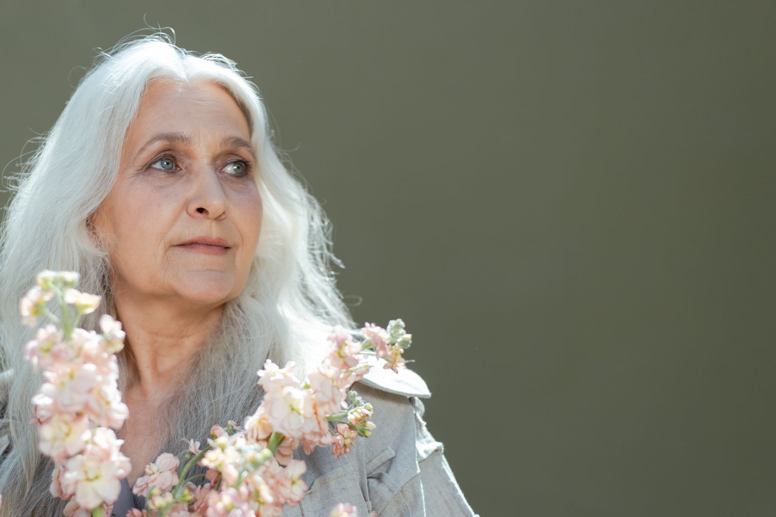 Woman over 60 with white hair looking away thoughtfully, holding soft pink flowers against a muted green background