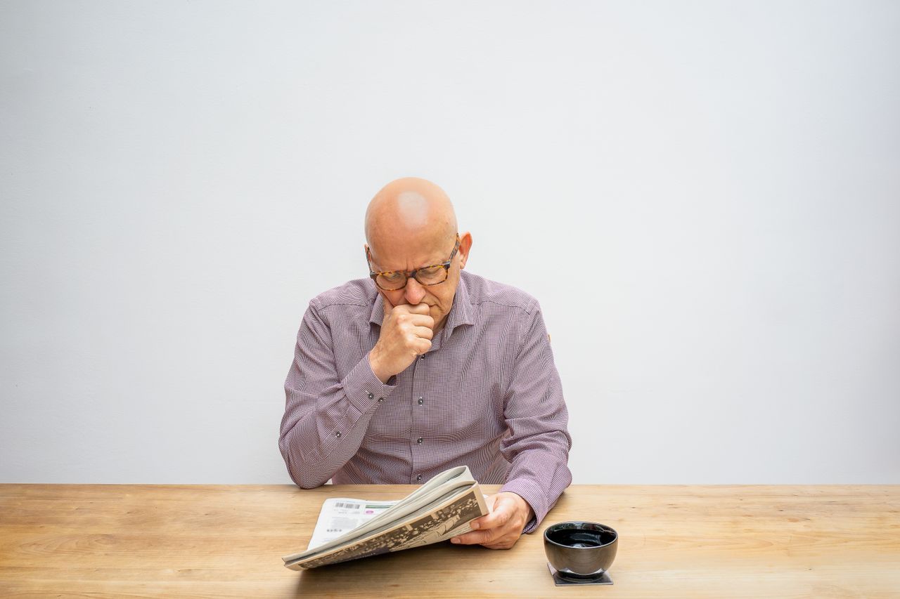 Bald man in glasses reading a newspaper at a wooden table with a cup of coffee