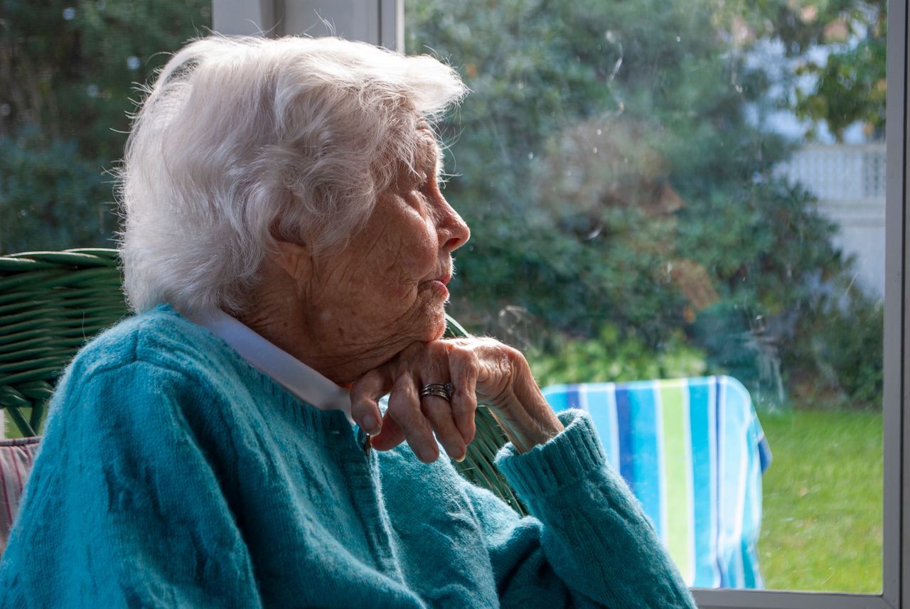 Senior woman with white hair leaning on her hand near a window at home, looking pensive