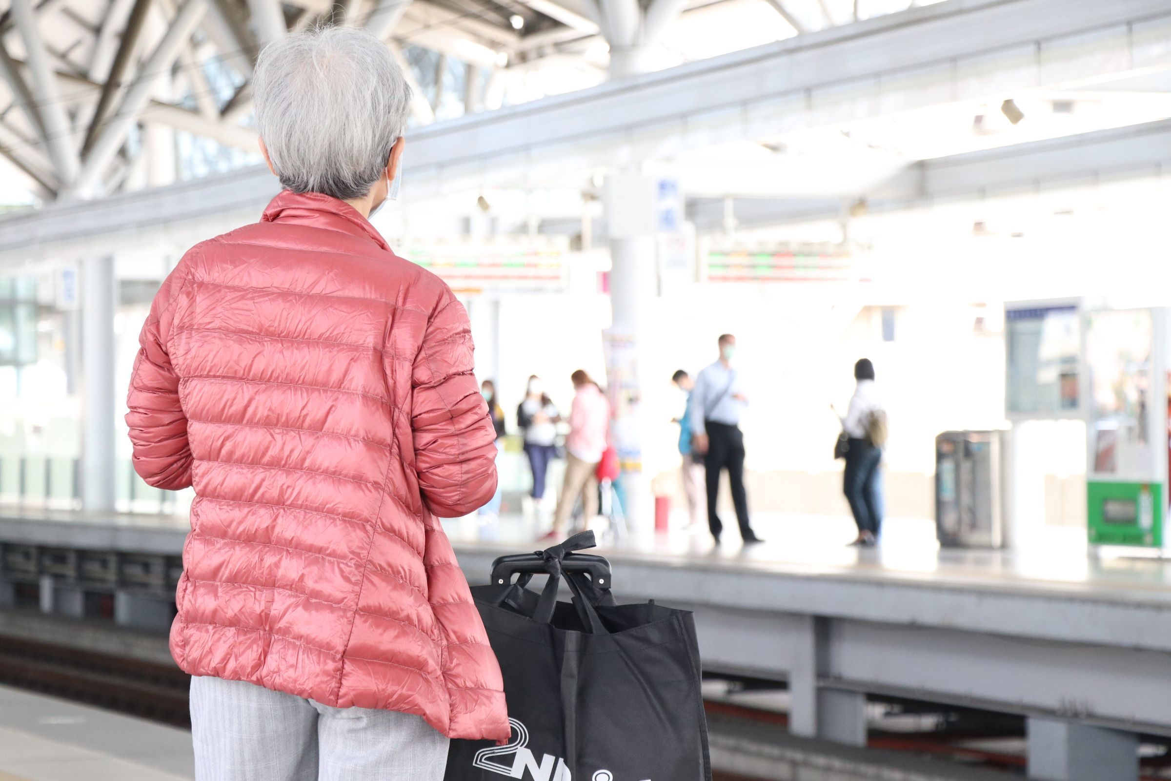 Elderly woman in a jacket waiting at a train station platform with luggage on a busy morning