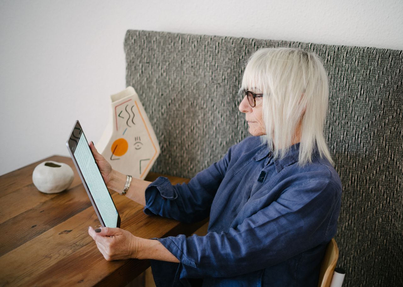 Senior woman with white hair and glasses reading a tablet at a wooden table at home