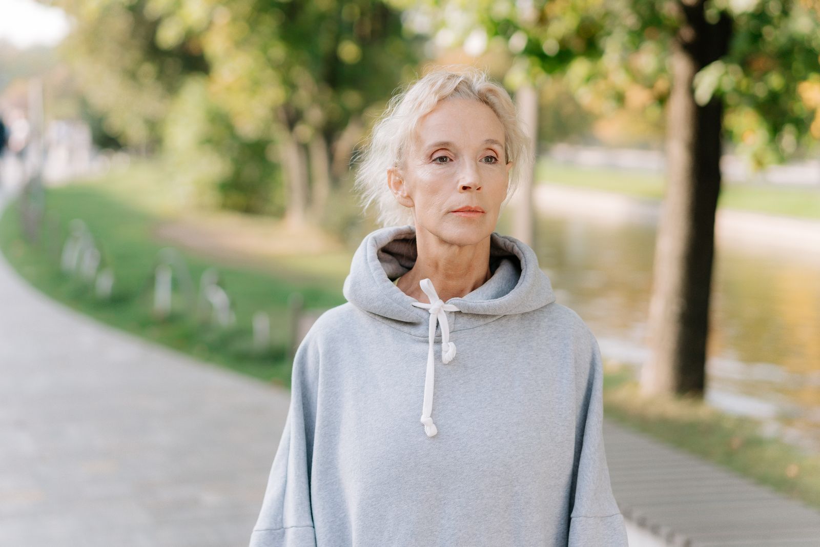 Mature woman with white hair standing on a tree-lined park path in a gray hoodie