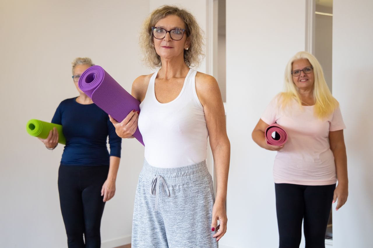 Three senior women holding yoga mats and smiling as they arrive for a class together