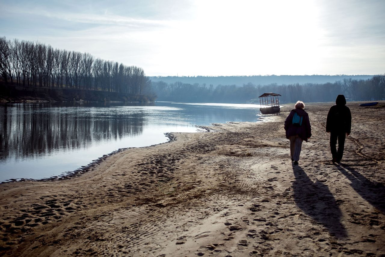 Two people walking side by side along a quiet riverbank in soft winter light