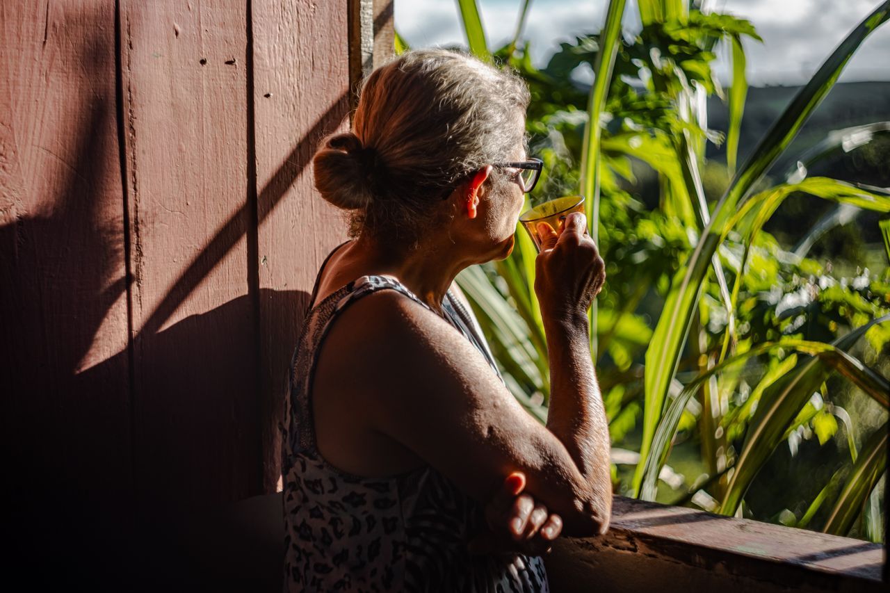 Senior woman with coffee on a sunny balcony surrounded by green plants, quiet morning alone