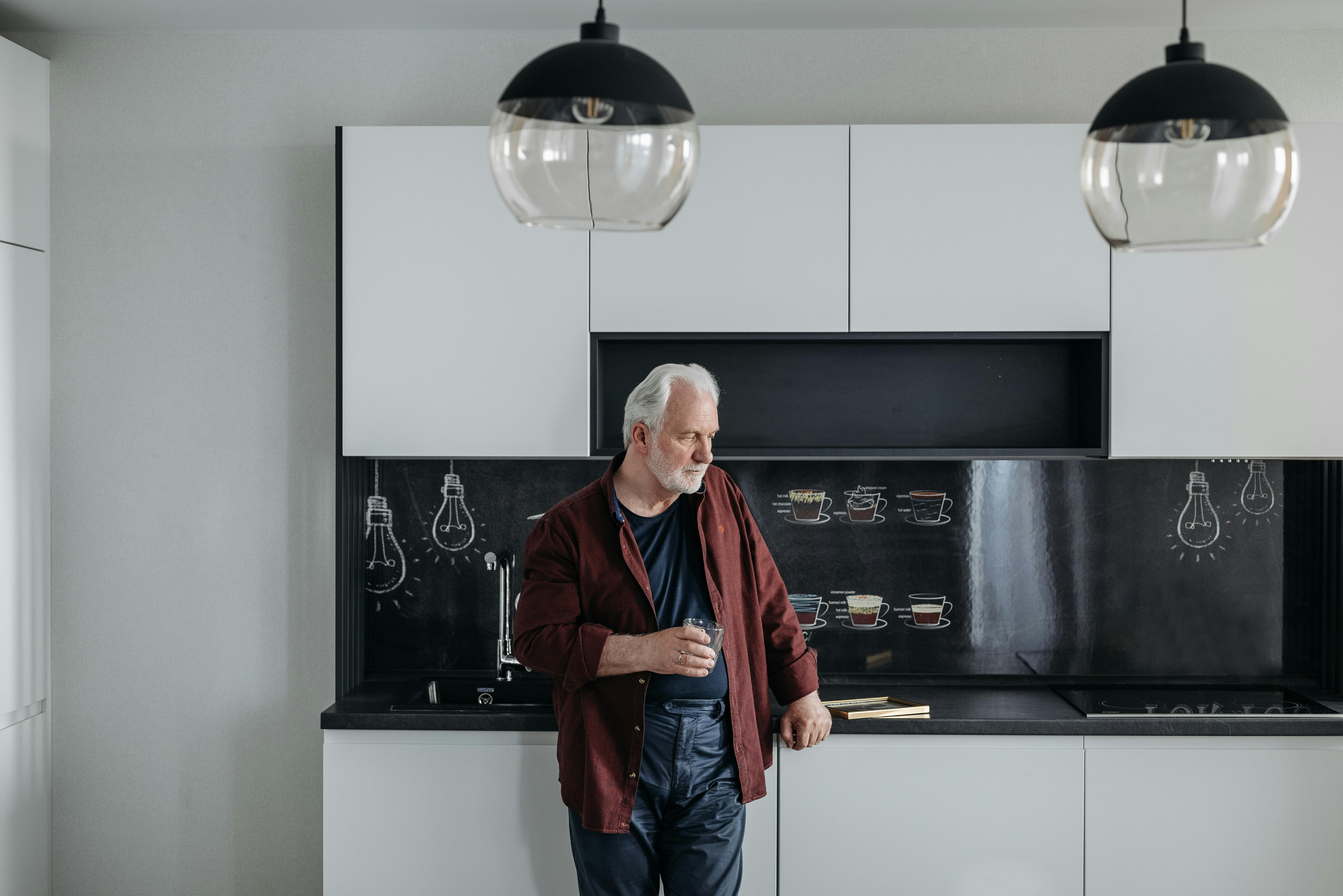 Senior man standing alone in a kitchen holding a glass, looking down in thought