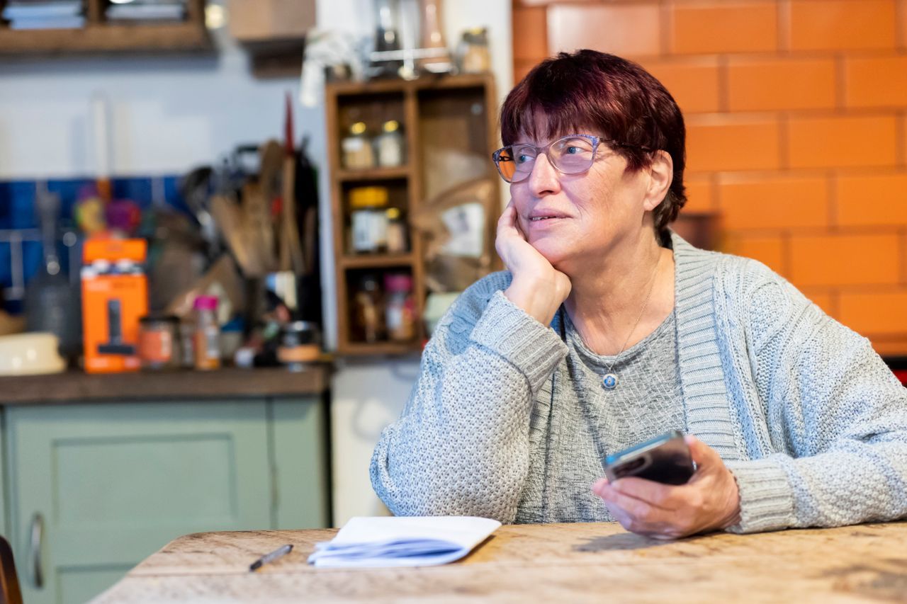 Older woman with glasses sitting at a kitchen table holding her phone and looking away thoughtfully