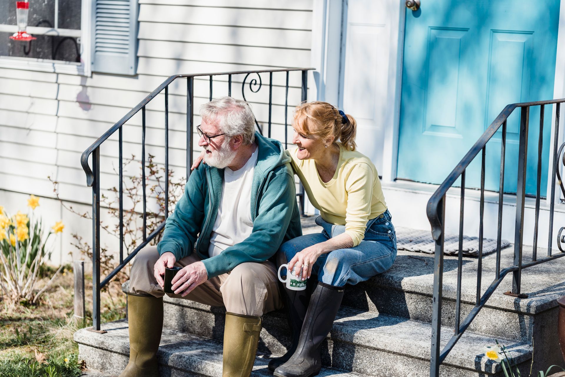 Older couple sitting together on porch steps with mugs looking out at their garden