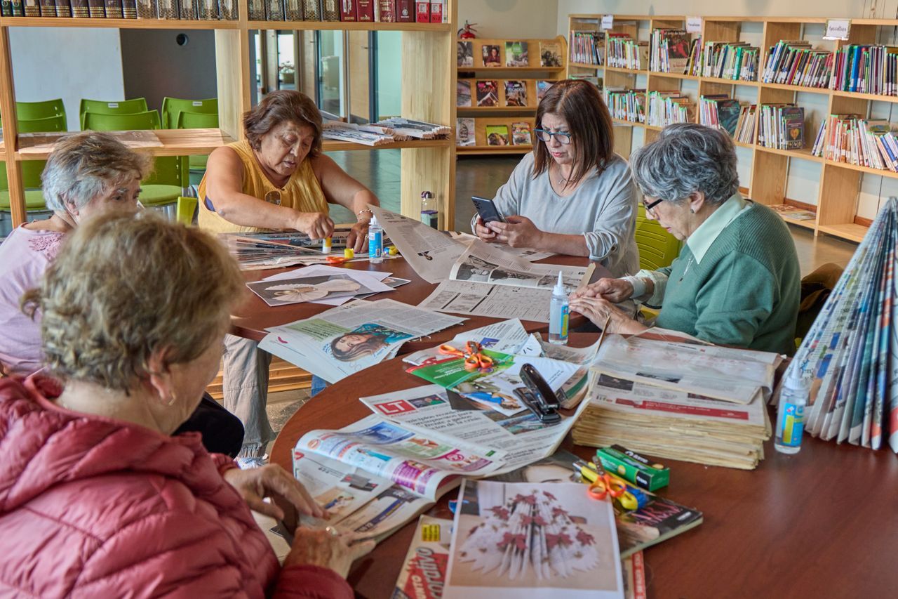 Older women working on a craft project at a library table with newspapers and scissors spread between them