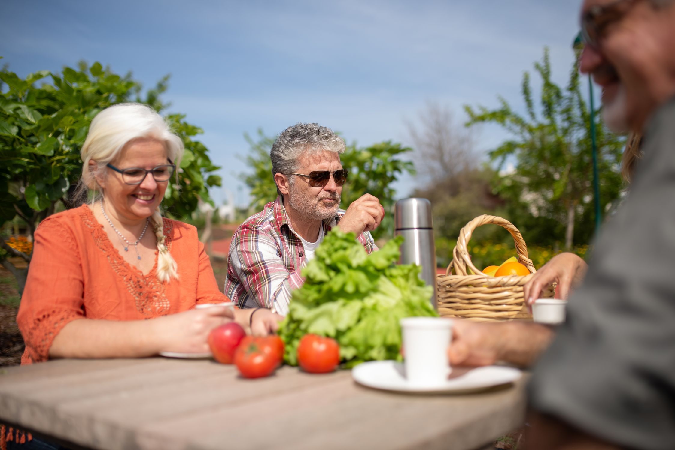 Older adults sharing coffee and fresh produce at an outdoor community garden table in warm sunshine