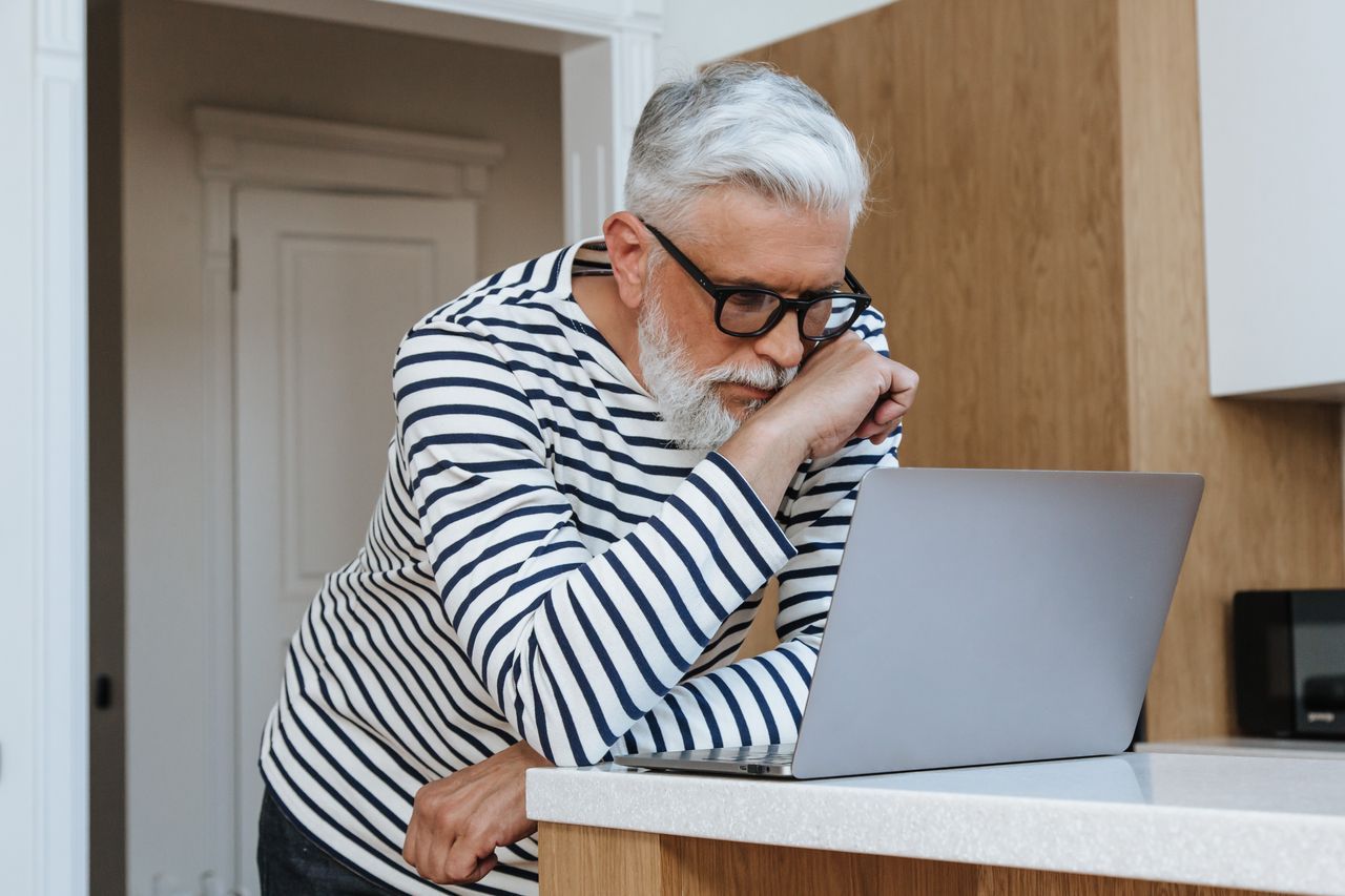Older man reviewing photos on a laptop at a kitchen table in morning light