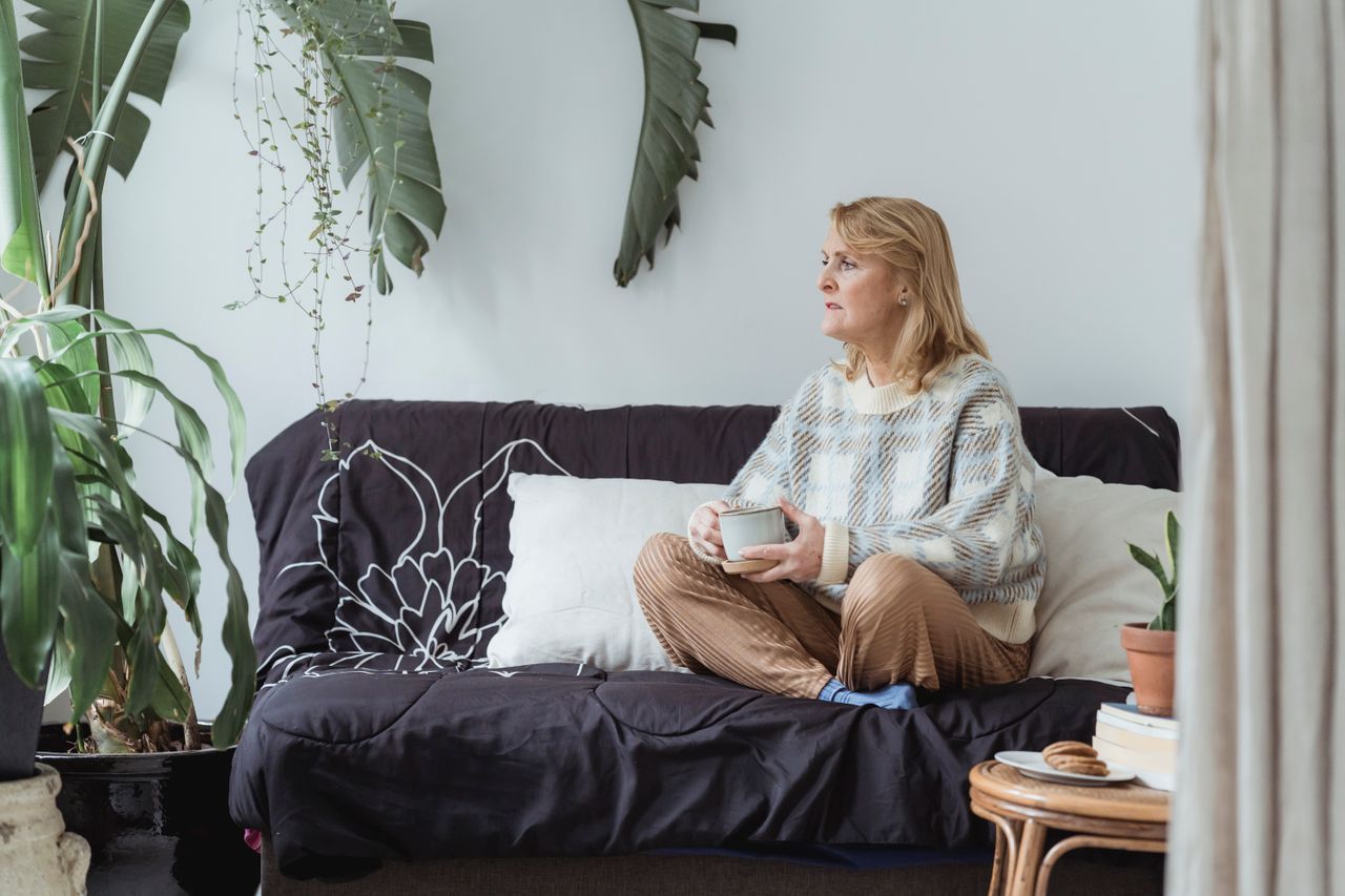 Mature woman with crossed legs drinking tea thoughtfully on a sofa at home