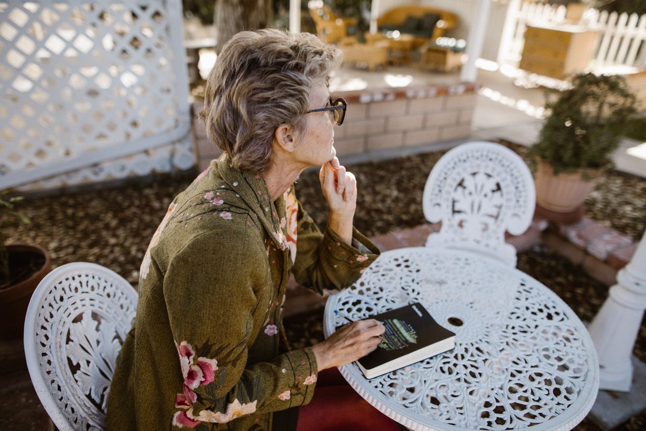 Older woman with glasses sitting thoughtfully at a garden patio table with a book