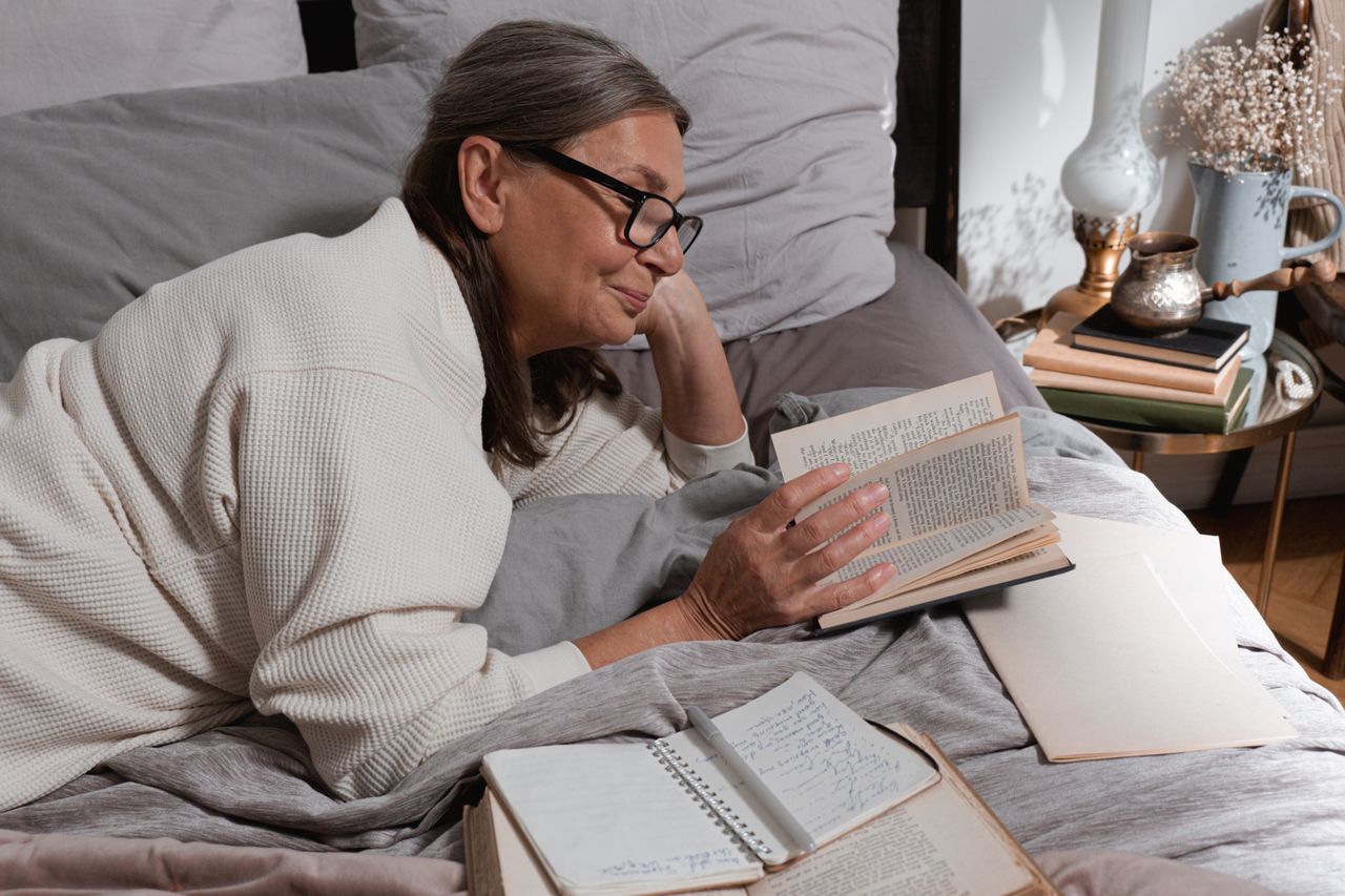 Mature woman with glasses reading a book in bed surrounded by journals and warm lamplight
