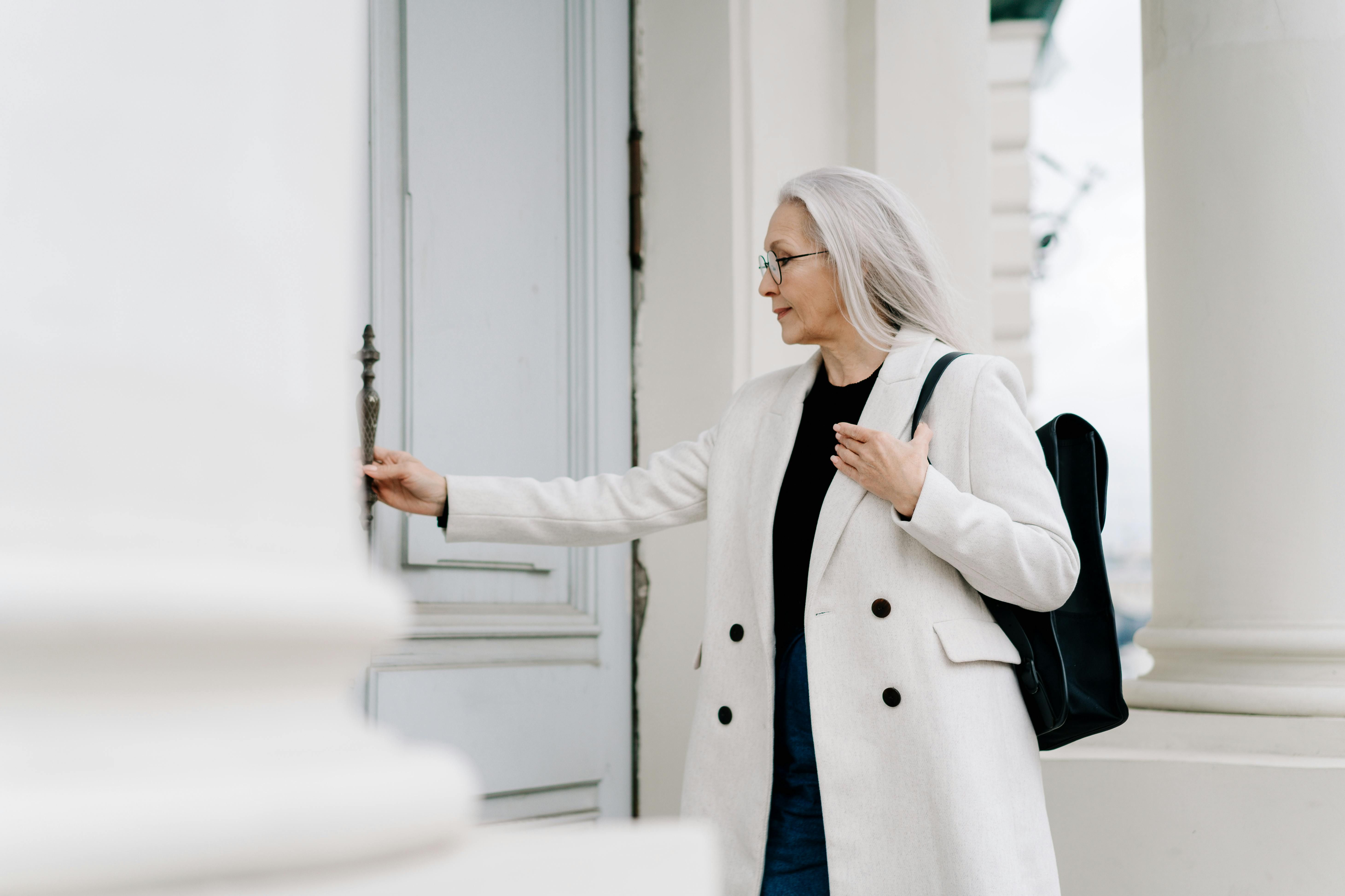 Silver-haired woman in white coat opening a door at a columned entrance