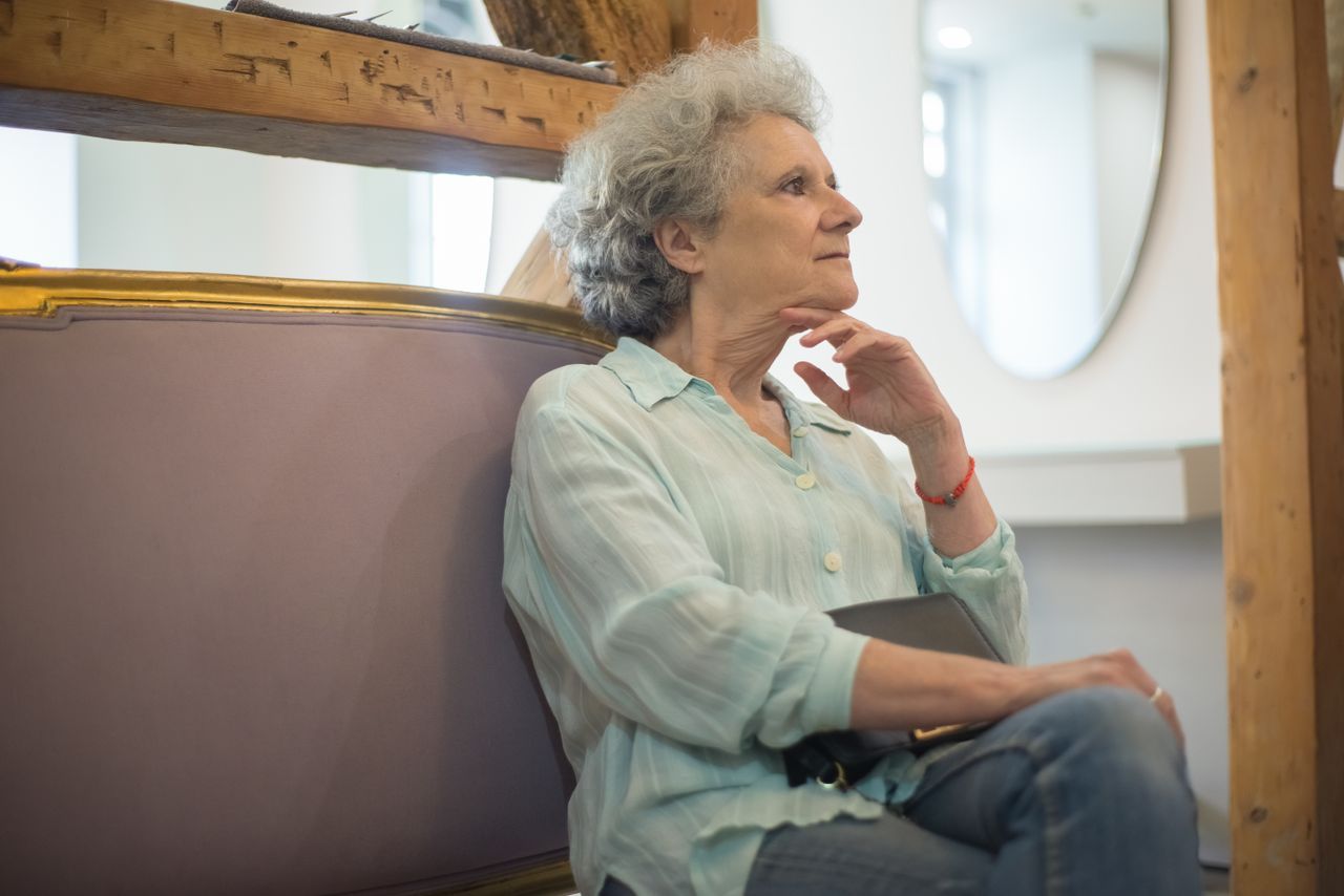 Grey-haired woman sitting on a sofa with hand on chin looking thoughtfully to the side