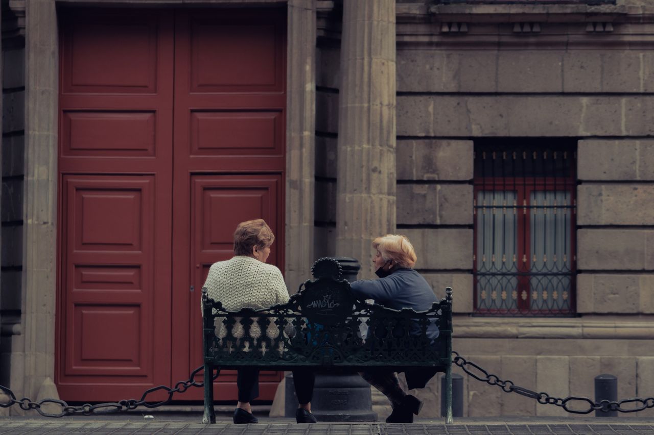Two older women sitting together on a bench outside a stone building, talking in warm afternoon light