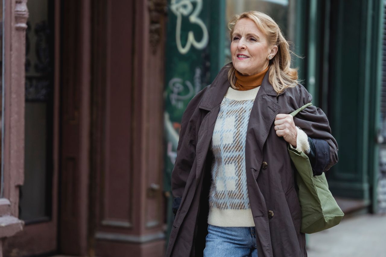 Mature woman in a checkered coat walking along a city sidewalk, looking away thoughtfully