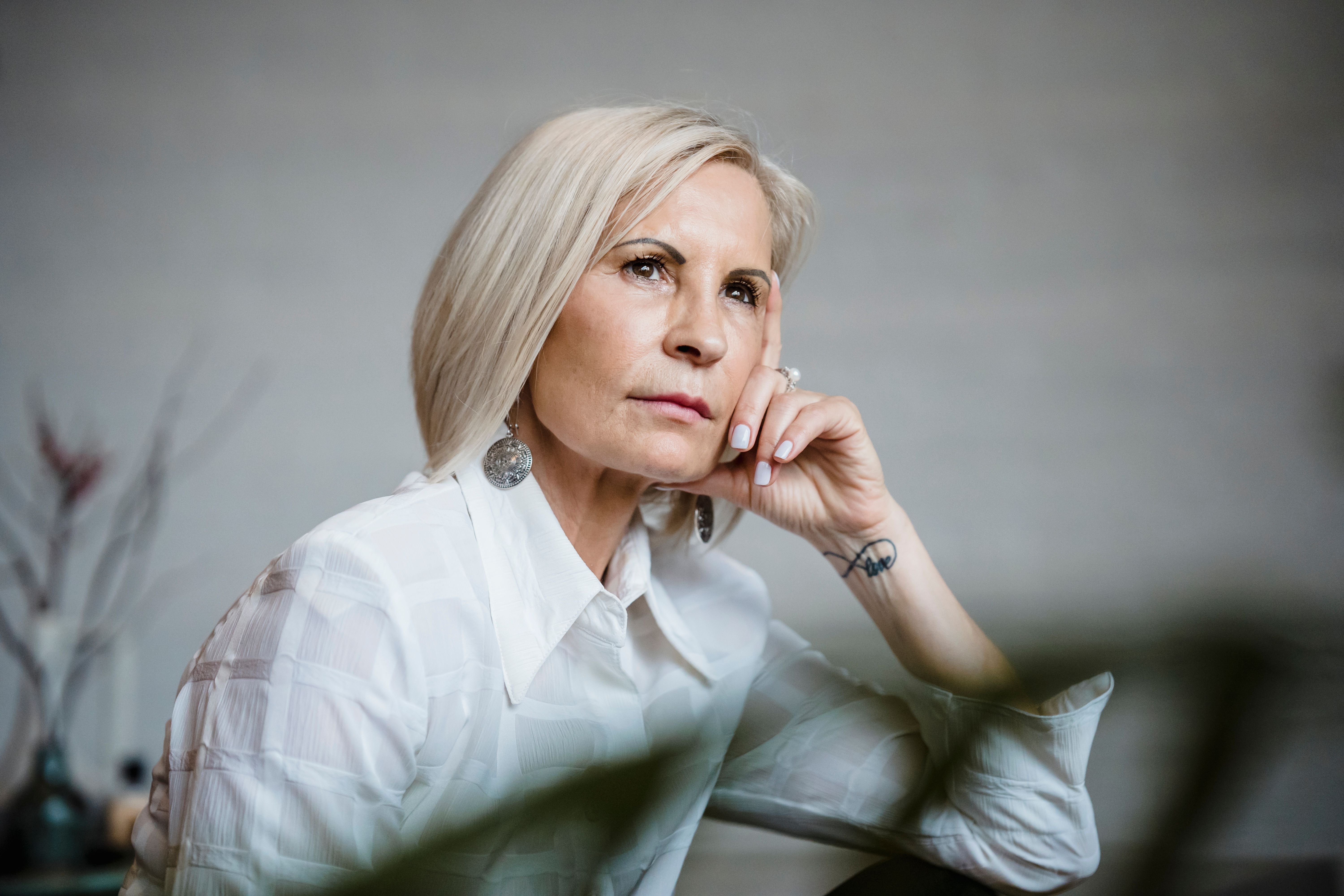Mature woman with platinum hair resting chin on hand in quiet thought, indoors in natural light