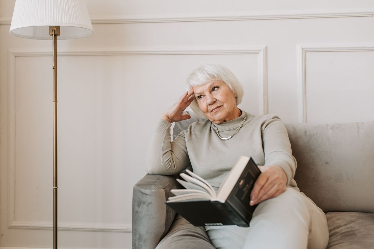 White-haired woman over 60 sitting on a sofa with a book, hand at her temple, looking thoughtful