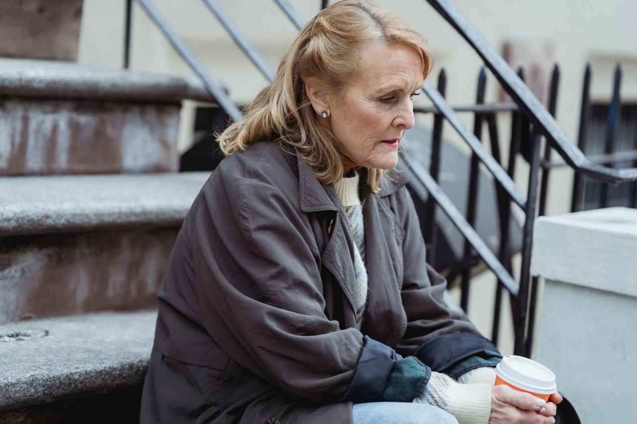 Senior woman in outerwear sitting on stone steps outdoors, looking down reflectively