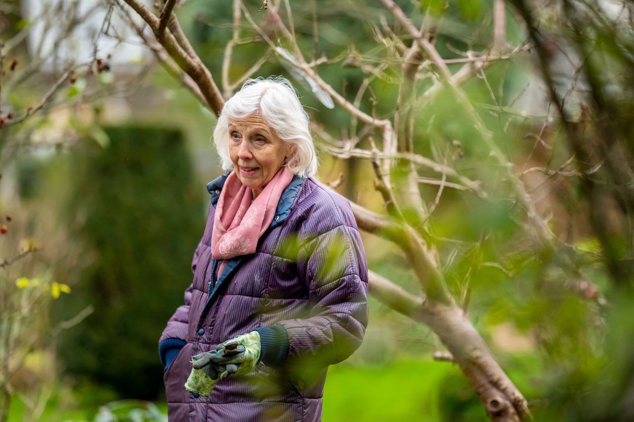 Older woman standing alone in a garden, looking thoughtful among bare branches
