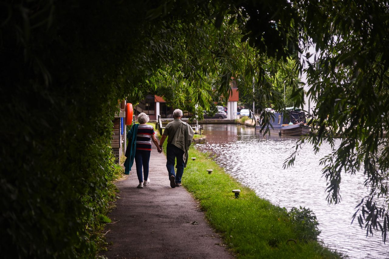 Older couple walking hand in hand along a canal path seen from behind through a leafy archway